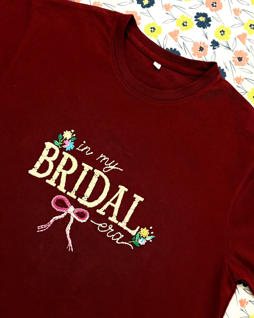 In My Bridal Era hand embroidered maroon tee laid flat on a floral print – cream lettering, pink bow, wildflowers