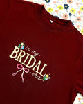 In My Bridal Era hand embroidered maroon tee laid flat on a floral print – cream lettering, pink bow, wildflowers